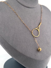 Load and play video in Gallery viewer, The newest design by Adeline - this unusual necklace is a tennis lovers delight.   The solid gold ball threads through the racket and can be worn higher or lower on the neck.  This piece was hand made in the jewelry district of Los Angeles by a fine jewel craft team.  The racket is 1.5&quot; long and the ball is 6mm around.  Light yet strong enough to play (and win) in.
